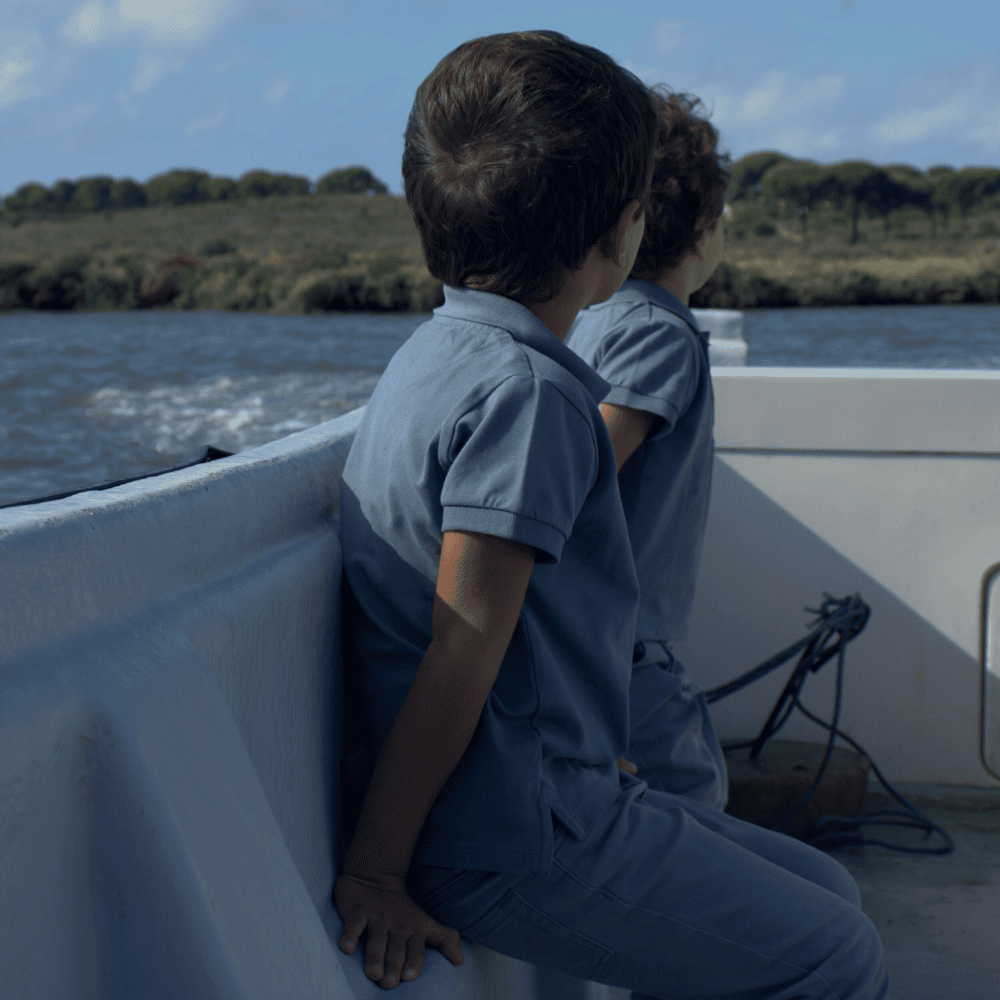 ABOUT_ Two children sitting on the edge of a boat, facing away, while looking out at the water and surrounding landscape.